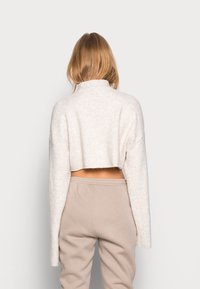 Geknipte, crèmekleurige trui met een hoge kraag en lange mouwen, gemaakt van een gestructureerde stof. Gecombineerd met beige joggingsbroek.