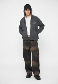 Šedá mikina na zip s předními kapsami, bílé tričko a hnědé maskáčové cargo kalhoty. Černé tenisky a ladící kšiltovka doplňují outfit.