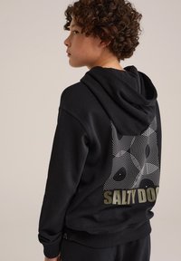 Melna kapuce ar teksturētu apdari, kurai ir balta viļņu raksts un uz muguras bolda olīvu krāsā rakstīts "SALTY DOG".