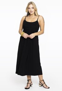 Schwarzes Maxikleid mit verstellbaren Spaghettiträgern, geraffter Taille, fließendem Rock und kombiniert mit verzierten schwarzen Sandalen.