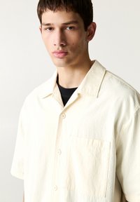 Camicia a maniche corte con colletto in tessuto bianco crema con finitura testurizzata, dotata di taschino sul petto e bottoni di colore chiaro.