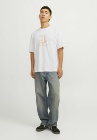 Witte katoenen T-shirt met een roze graphic met de tekst "On The Go." Gecombineerd met lichtblauwe baggy jeans en zwarte schoenen.