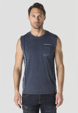 Hombre vistiendo una camiseta sin mangas azul marino con bolsillo y texto, combinada con vaqueros oscuros, de pie frente a un fondo claro y liso.