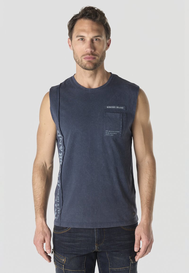 Hombre vistiendo una camiseta sin mangas azul marino con bolsillo y texto, combinada con vaqueros oscuros, de pie frente a un fondo claro y liso.