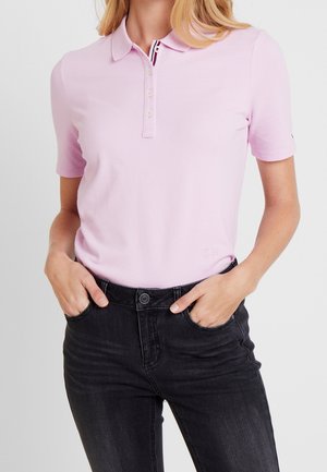 Frau trägt ein hellrosa Poloshirt mit kurzen Ärmeln, das in eine hoch taillierte schwarze Jeans gesteckt ist, mit den Händen in den vorderen Taschen.