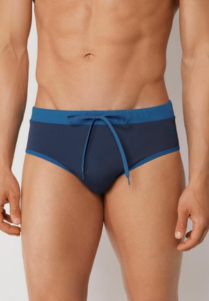 Männlicher Oberkörper mit marineblauen Badehosen mit einem hellblauen Bund und vorne zu einer Schleife gebundenem Kordelzug.