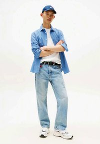 Mlad moški z ovitimi rokami, oblečen v modro karirasto srajco, belo majico, svetle kavbojke, črni pas, bele superge in modro kapo za bejzbol.