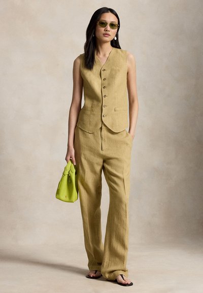Γυναίκα με μουσταρδί κίτρινη αμάνικη γιλέκο με κουμπιά και φαρδιά παντελόνια, κρατώντας μια τσάντα σε lime πράσινο χρώμα και φοράει γυαλιά ηλίου.