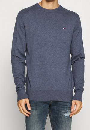 Mann trägt einen schlichten dunkelgrauen Pullover mit rundem Ausschnitt und ausgewaschene blaue Jeans mit einem Flicken am linken Oberschenkel, steht vor einem neutralen Hintergrund.