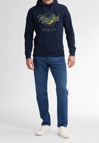 Námořnicky modrá mikina s kapucí s grafickým potiskem v světle zelené barvě, vybavená klokankou, kapucí se šňůrkami a žebrovanými manžetami. Kombinována s tmavě modrými džínami a bílými teniskami.