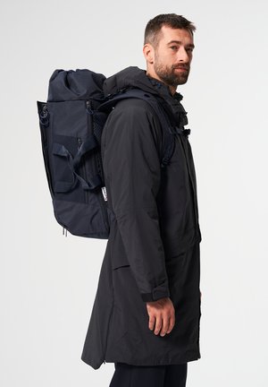 Mann trägt eine lange schwarze Jacke und einen großen dunkelblauen Rucksack, steht im Profil vor einem einfarbigen Hintergrund.