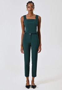Dunkelgrüner Jumpsuit mit quadratischem Ausschnitt, tailliertem Oberteil und schmal zulaufenden Beinen, ausgestattet mit einem Stoffgürtel mit einer runden Schnalle.