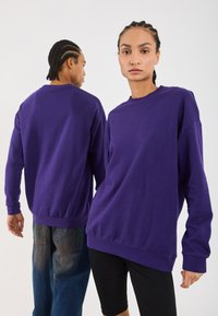 Twee mensen met gevlochten haar die paarse sweatshirts dragen, waarvan de een naar voren kijkt en de ander zich omdraait, tegen een effen witte achtergrond.
