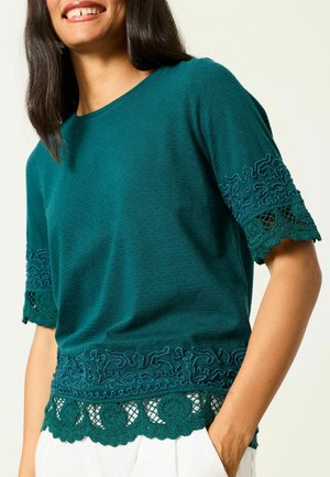 Woman wearing teal short-sleeve top with embroidered and lace details on sleeves and hem, paired with white pants, smiling.