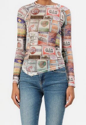 Kvinde iført en tætsiddende langærmet top med farverige vintage-inspirerede benzinskilte og racermærke-patches, kombineret med blå cowboybukser.