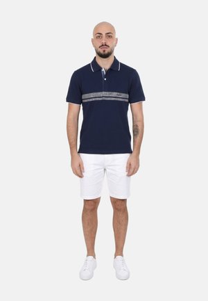 Uomo in piedi di fronte, che indossa una polo blu navy con una striscia grigia, pantaloncini bianchi, scarpe da ginnastica bianche e un tatuaggio sull'avambraccio sinistro.