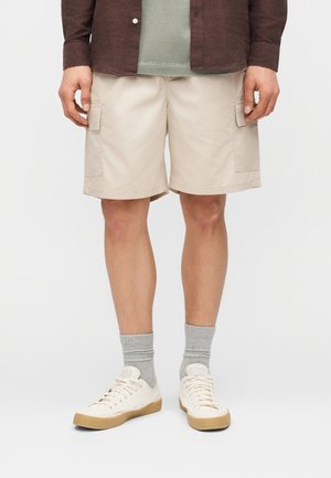 Mężczyzna w beżowych szortach typu cargo, szarych skarpetkach, białych trampkach, zielonej koszulce i brązowej, rozpiętej kurtce, stojący na białym tle.