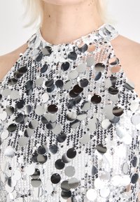 Silver sequin halter top featuring circular sequins on a sheer fabric with a high neckline and open shoulder design. Textured and reflective.