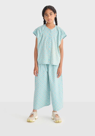 Fille aux cheveux tressés portant un haut boutonné bleu clair à motifs et un pantalon large, debout avec une expression neutre et des chaussures beiges.