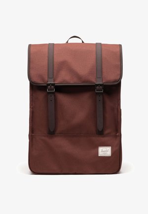 Brown fabric backpack with a flap closure, featuring two dark brown straps and a front pocket. Label on the bottom right corner.