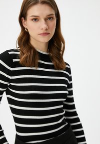 Raitapintainen mustavalkoinen ribattua pitkähihainen paita, jossa on pyöreä pääntie. Ominaisuuksiin kuuluu teksturoitu kangas, vartalonmyötäinen malli ja kontrastivärinen trimmi pääntien ympärillä.