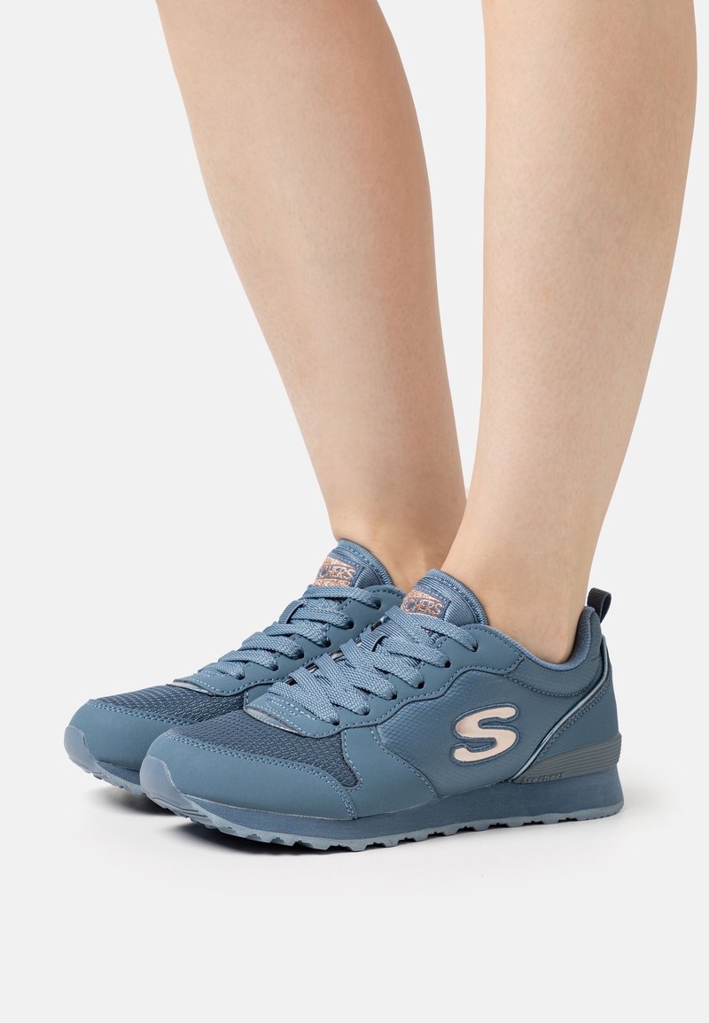 Baskets Skechers bleues avec panneaux en mesh et cuir, design à lacets et logo rose "S", portées pieds nus sur fond blanc.