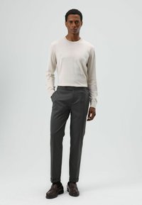 Beige lange mouw trui en op maat gemaakte donkergrijze broek. Schoenen zijn bruine loafers. Model staat met handen in de zakken tegen een neutrale achtergrond.