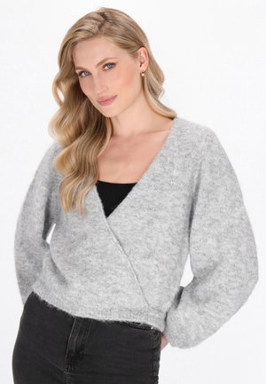 Donna bionda con capelli mossi che indossa orecchini a cerchio dorati, un morbido maglione grigio avvolgente sopra un top nero e jeans neri a vita alta.