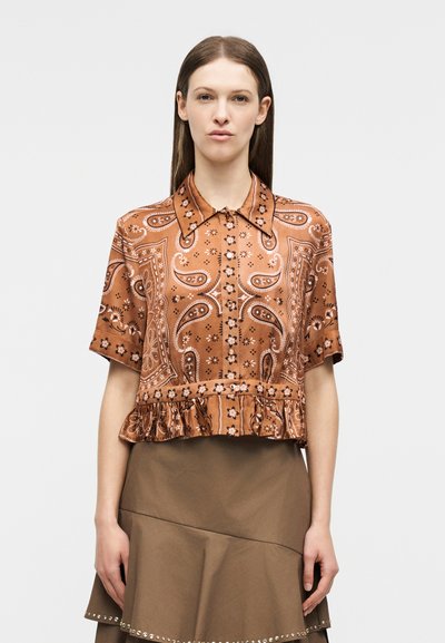 Νεαρή γυναίκα με ίσια καστανά μαλλιά, που φοράει μια γυαλιστερή καφέ μπλούζα με κοντά μανίκια και σχέδιο, και μια καφέ φούστα με βολάν και τρουκς.