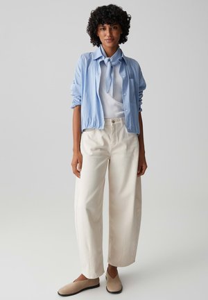 Vrouw met krullend haar die een lichtblauw gestreept shirt, een wit topje, beige brede broek en beige instapschoenen draagt, staand tegen een effen achtergrond.