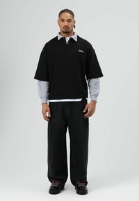 Zwart oversized poloshirt met grijze accenten, gedragen over een wit shirt, gecombineerd met losse zwarte spijkerbroek en grove zwarte schoenen.