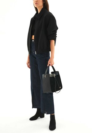 Giacca bomber nera con una silhouette aderente, abbinata a jeans in denim scuro a gamba larga. Tiene una borsa nera texturizzata con hardware dorato.