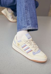 Zapatillas beige con rayas azul claro y rojas, con detalles de ante, material texturizado y suela de goma. Logotipo dorado en el lado.