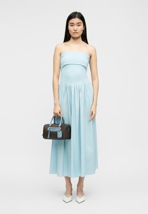 Γυναίκα με αμάνικο, ανοιχτό μπλε φόρεμα μήκους μέχρι τη μέση της γάμπας, κρατώντας μια μικρή καφέ τσάντα με μπλε χερούλια και φορώντας άσπρα μυτερά τακούνια.