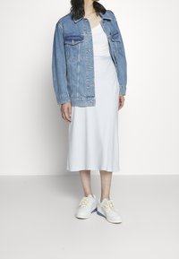 Džínová bunda, světle modrá, se dvěma kapsami na hrudi, vrstvená přes bílý tílko. Midi-článek, přiléhavé bílé šaty. Bílé tenisky s modrými akcenty.