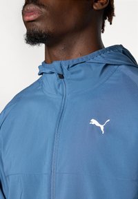 Plava sportska jakna s patentnim zatvaračem sprijeda, kapuljačom i bijelim Puma logotipom na prsima. Materijal izgleda lagan i glatko.