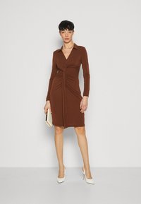 Robe marron à manches longues avec un décolleté en V, présentant des détails froncés à la taille et une coupe ajustée. Associée à des chaussures à talons blancs.