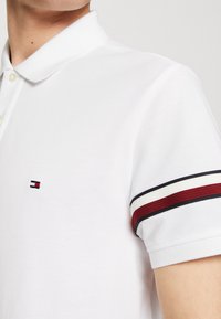 Vit pikétröja i texturerat tyg, med en krage med tre knappar och röd-, vit- och marinblå randiga detaljer på ärmarna, med logotyp.