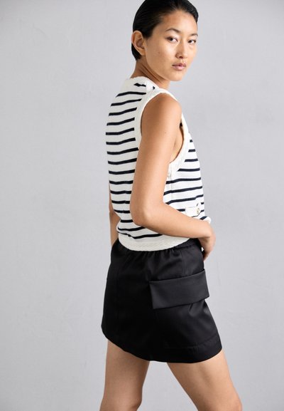 Ριγέ αμάνικο τοπ σε λευκό και ναυτικό χρώμα, σε συνδυασμό με μια μαύρη σατέν φούστα που διαθέτει πλάγια τσέπη. Το μοντέλο ποζάρει από γωνία τριών τετάρτων.