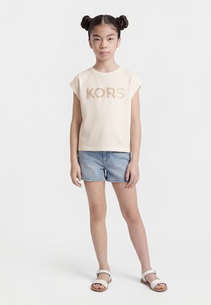 Young girl with double buns wearing a beige KORS T-shirt, denim shorts, and white sandals standing against a plain white background.