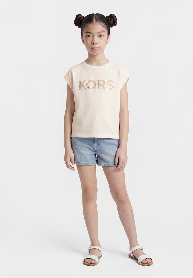 Jeune fille avec des double chignons portant un T-shirt beige KORS, un short en jean et des sandales blanches, debout devant un fond blanc uni.