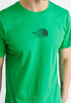 Mężczyzna w jasnooliwkowej koszulce z krótkim rękawem z czarnym logo i napisem "The North Face" na lewej piersi.