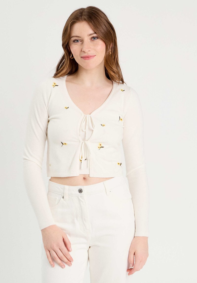 Cream-colored cropped cardigan with long sleeves, V-neck, and tie front. Features yellow floral embroidery for decoration.