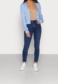 Ljusblå ribbade cardigan med långa ärmar, beige topp med paljettdetaljer och mörkblå skinny jeans, matchat med vita sneakers.