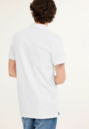 Mann mit lockigem braunem Haar, der ein einfaches weißes Polohemd mit kurzen Ärmeln und blaue Jeans trägt und ein Sonnen-Tattoo auf seinem rechten Arm zeigt.