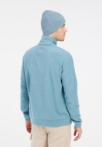 Šviesiai mėlynas poliarinės vilnos megztinis su aukšta apykakle, ilgomis rankovėmis ir lygia tekstūra. Derinamas su raukšlėtu šviesiai mėlynu kepurėliu.