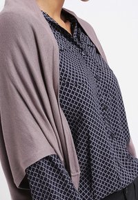 Leichtes, fliederfarbenes Tuch, das über einer marineblauen Bluse mit einem geometrischen weißen Muster drapiert ist. Die Bluse verfügt über eine durchgehende Knopfleiste und einen Kragen.