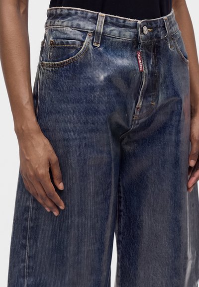 Gros plan d'une personne portant un jean bleu délavé à jambes larges avec une étiquette rouge Dsquared2 près de la poche avant et les mains détendues sur les côtés.