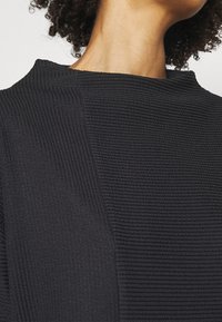 Top nero a coste con colletto alto e tessuto texturizzato. Il design include un pannello liscio ai lati per creare contrasto.