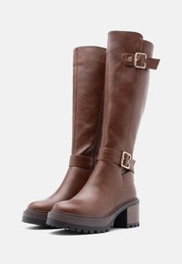 Brown leather knee-high boots with a side zipper, featuring a buckle accent and a chunky textured sole. Stacked heel for added height.